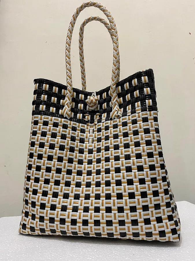 Foto asli: tas anyaman plastik bermotif kotak hitam, putih, dan emas dengan tali anyaman, difoto agak menyamping di depan dinding krem