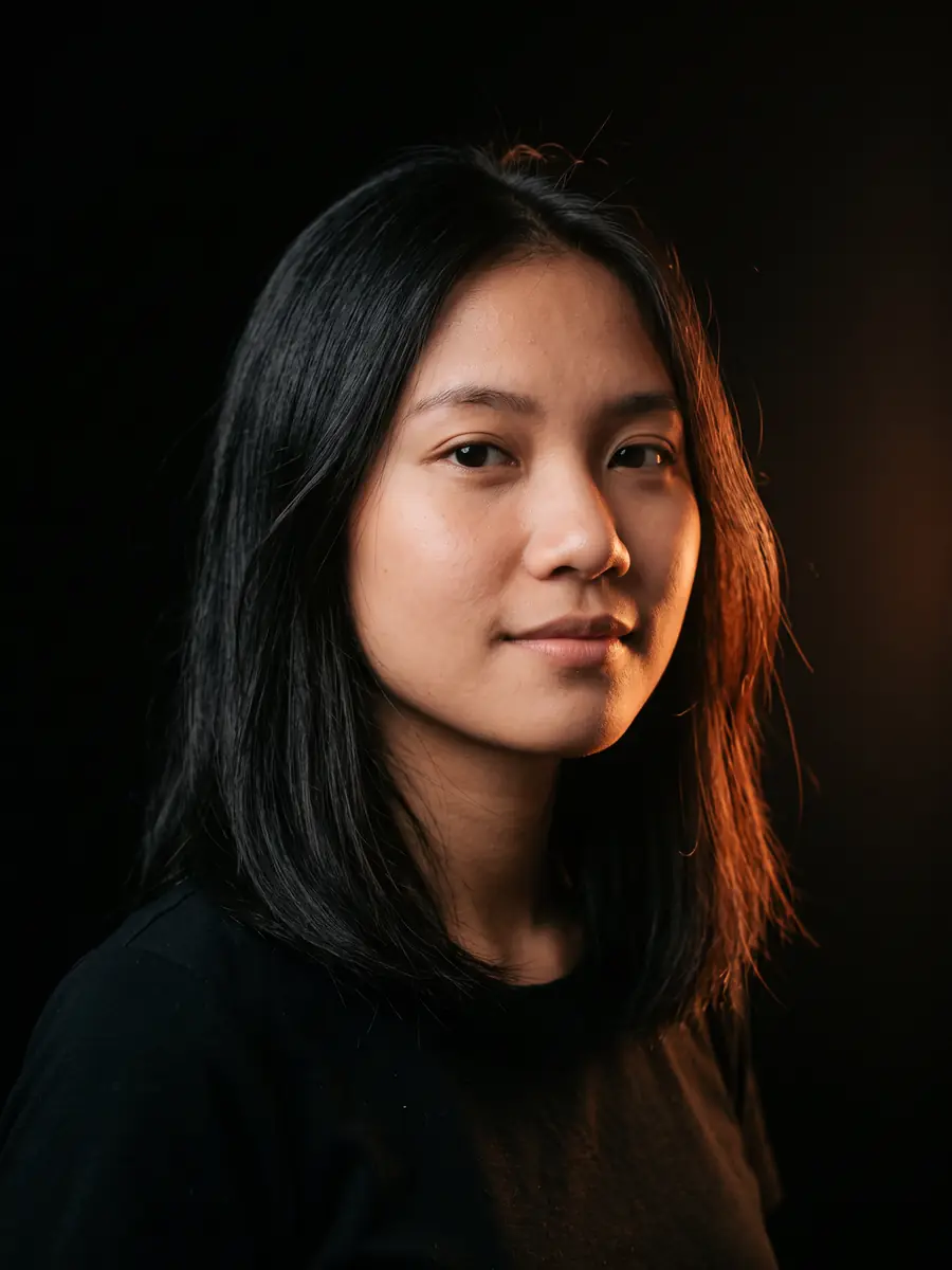 Potret perempuan muda Asia Tenggara fiktif berambut hitam sebahu dan berbaju hitam, latar hitam, cahaya hangat dari samping kanan.