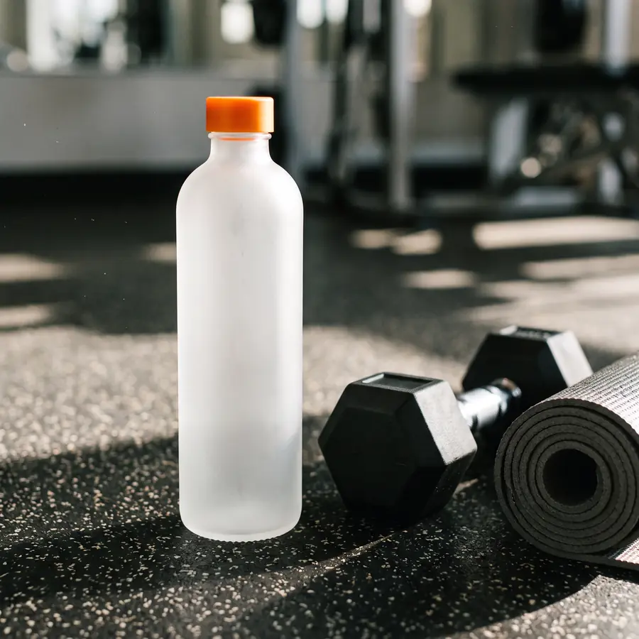 Botol minum kaca buram bertutup jingga tanpa label di lantai karet gym di samping dumbel hitam dan matras yoga tergulung.