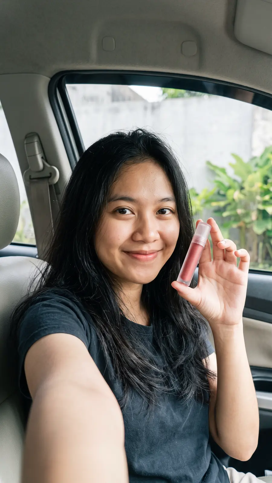 Kreator fiktif perempuan muda berambut panjang berswafoto di dalam mobil sambil menunjukkan tube lip tint tanpa label.